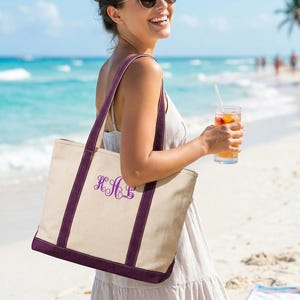 May include: A beige canvas tote bag with purple trim and straps. The bag features a purple monogram and is being carried on a beach. The background includes the ocean, sand, and a person holding a drink.