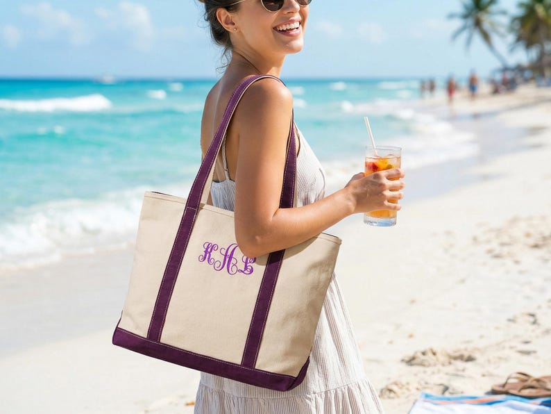 May include: A beige canvas tote bag with purple trim and straps. The bag features a purple monogram and is being carried on a beach. The background includes the ocean, sand, and a person holding a drink.
