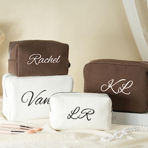 Puede incluir: Cuatro neceseres personalizados en marrón y blanco. Las bolsas rectangulares tienen cierre de cremallera y nombres o iniciales bordados en letra cursiva. Pinceles de maquillaje y perlas en primer plano.