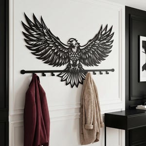 May include: A black metal wall art piece featuring a detailed eagle with outstretched wings. Below the eagle is a black coat rack with multiple hooks. A burgundy jacket and a beige cardigan hang from the hooks.