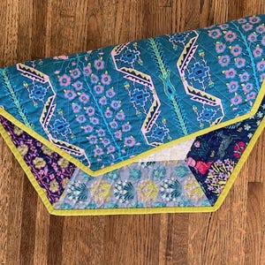 May include: A multi-colored quilted throw blanket with a geometric pattern and floral accents. The blanket has a yellow border and is made of fabric with a soft texture.