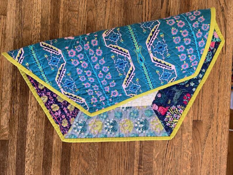 May include: A multi-colored quilted throw blanket with a geometric pattern and floral accents. The blanket has a yellow border and is made of fabric with a soft texture.