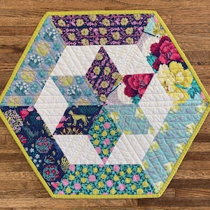 May include: A hexagonal table runner with a geometric pattern of white triangles and colorful floral fabric squares. The runner has a lime green border.