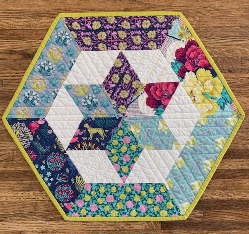 May include: A hexagonal table runner with a geometric pattern of white triangles and colorful floral fabric squares. The runner has a lime green border.