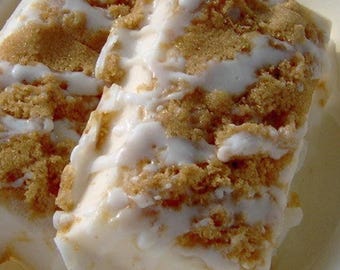 May include: Close-up of two rectangular desserts. The desserts have a creamy white base, topped with a layer of brown crumble and drizzled with a white glaze. The desserts are on a light yellow surface.