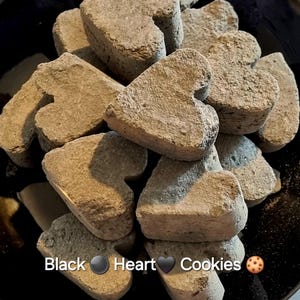 Peut inclure: Gros plan sur un tas de biscuits en forme de cœur gris foncé sur une assiette noire. Les biscuits sont disposés de manière aléatoire, certains se chevauchant. Le texte "Black Heart Cookies" est écrit sous les biscuits.