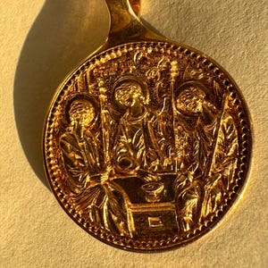 Op de afbeelding: Een goudkleurige hanger met een gedetailleerd reliëf van religieuze figuren. De hanger heeft een lus aan de bovenkant met een kruisontwerp. De figuren zijn afgebeeld in een cirkelvormig kader met ingewikkelde randdetails.