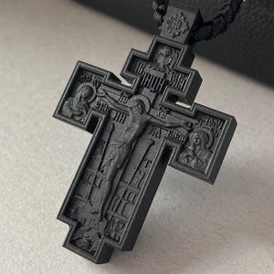 May include: A black Orthodox cross pendant with intricate carvings, suspended on a black cord necklace. The cross features a detailed depiction of the crucifixion and religious iconography. The necklace includes silver-toned metal accents.