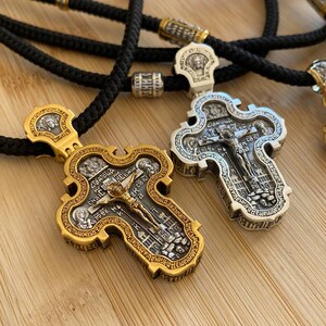 May include: Two ornate religious cross pendants with intricate detailing. One is gold-toned, the other silver-toned, both featuring religious figures and text. They hang on black cords with gold and silver accents, set against a light wooden surface.