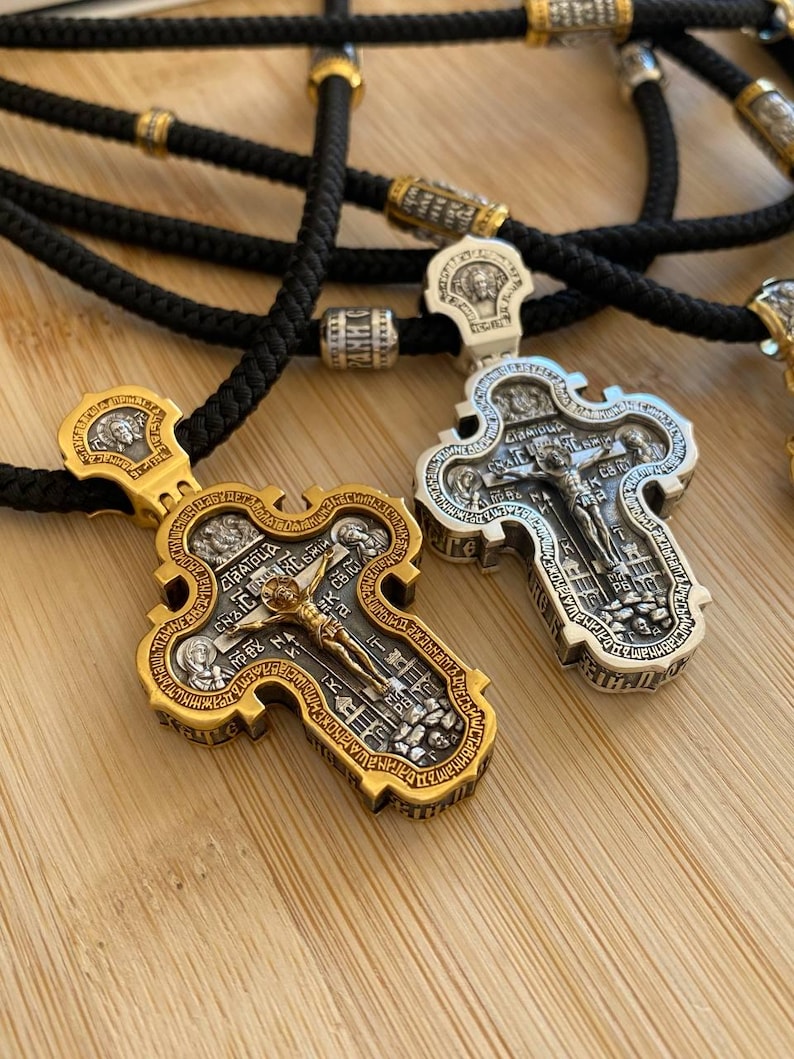 May include: Two ornate religious cross pendants with intricate detailing. One is gold-toned, the other silver-toned, both featuring religious figures and text. They hang on black cords with gold and silver accents, set against a light wooden surface.