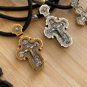 May include: Close-up of two ornate, gold and silver-toned Orthodox cross pendants with intricate detailing. The crosses feature a crucified Jesus figure and religious text. They are suspended on black cords, suggesting they are necklaces.