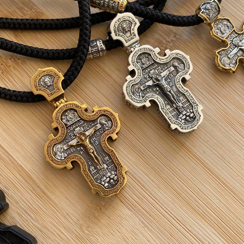May include: Close-up of two ornate, gold and silver-toned Orthodox cross pendants with intricate detailing. The crosses feature a crucified Jesus figure and religious text. They are suspended on black cords, suggesting they are necklaces.