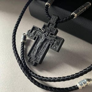 May include: A black Orthodox cross pendant with a detailed carved design, suspended from a braided black cord necklace. The cross features religious iconography and is accented with silver-toned metal details.