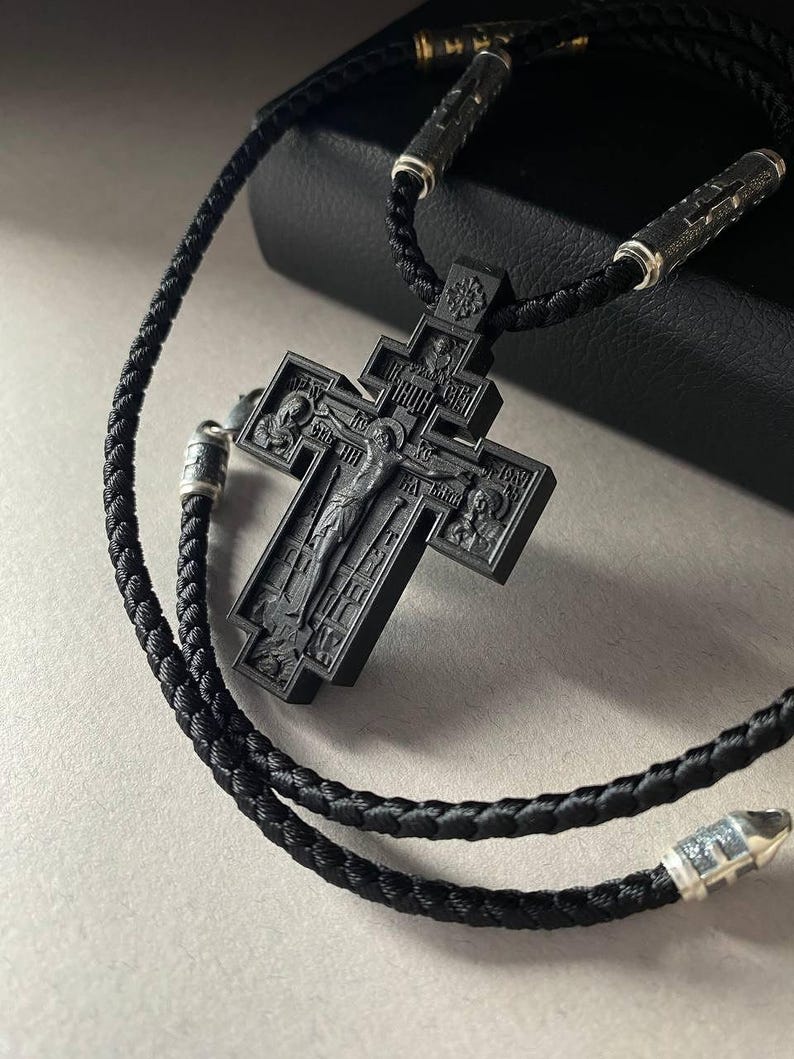 May include: A black Orthodox cross pendant with a detailed carved design, suspended from a braided black cord necklace. The cross features religious iconography and is accented with silver-toned metal details.