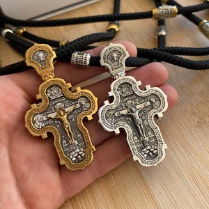 May include: Two ornate Orthodox Christian crosses, one gold-toned and one silver-toned, are displayed. Each cross features intricate religious iconography and a depiction of Jesus Christ. The crosses are suspended on black cords with decorative beads.