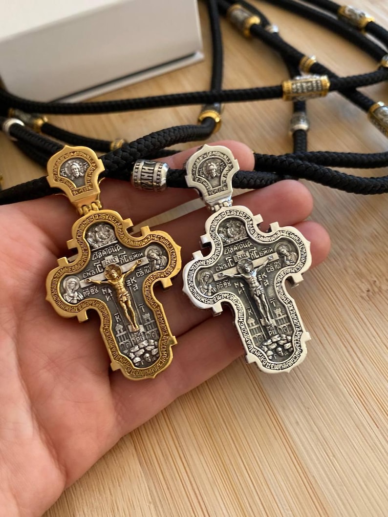 May include: Two ornate Orthodox Christian crosses, one gold-toned and one silver-toned, are displayed. Each cross features intricate religious iconography and a depiction of Jesus Christ. The crosses are suspended on black cords with decorative beads.
