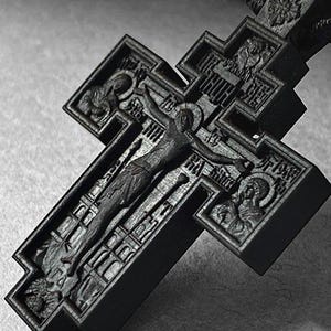 May include: A detailed, dark gray wooden Orthodox cross with intricate carvings. The cross features a central depiction of the crucifixion, surrounded by religious figures and inscriptions. The cross is suspended from a dark cord.