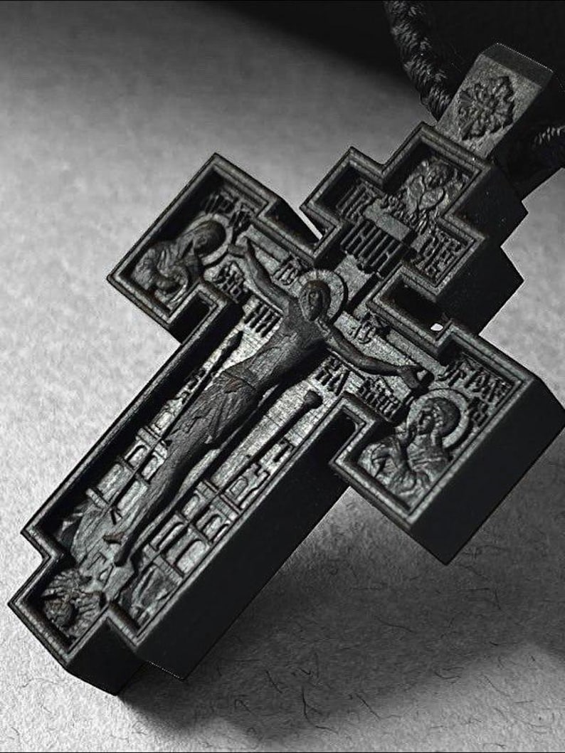 May include: A detailed, dark gray wooden Orthodox cross with intricate carvings. The cross features a central depiction of the crucifixion, surrounded by religious figures and inscriptions. The cross is suspended from a dark cord.