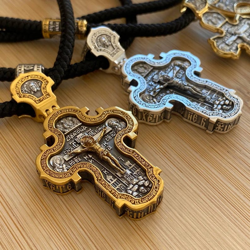 May include: Close-up of two ornate gold and silver religious cross pendants on black cords. The crosses feature detailed engravings of religious figures and text. The background is a light brown wooden surface.
