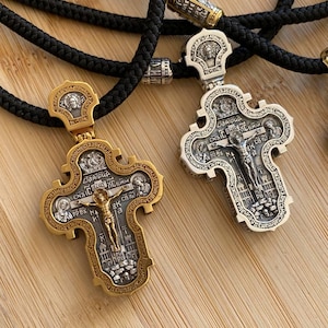 May include: Two ornate religious pendant necklaces. One is gold-toned, the other silver-toned, both with intricate detailing and a central figure. The pendants are suspended from black braided cords. The background is a light wood surface.