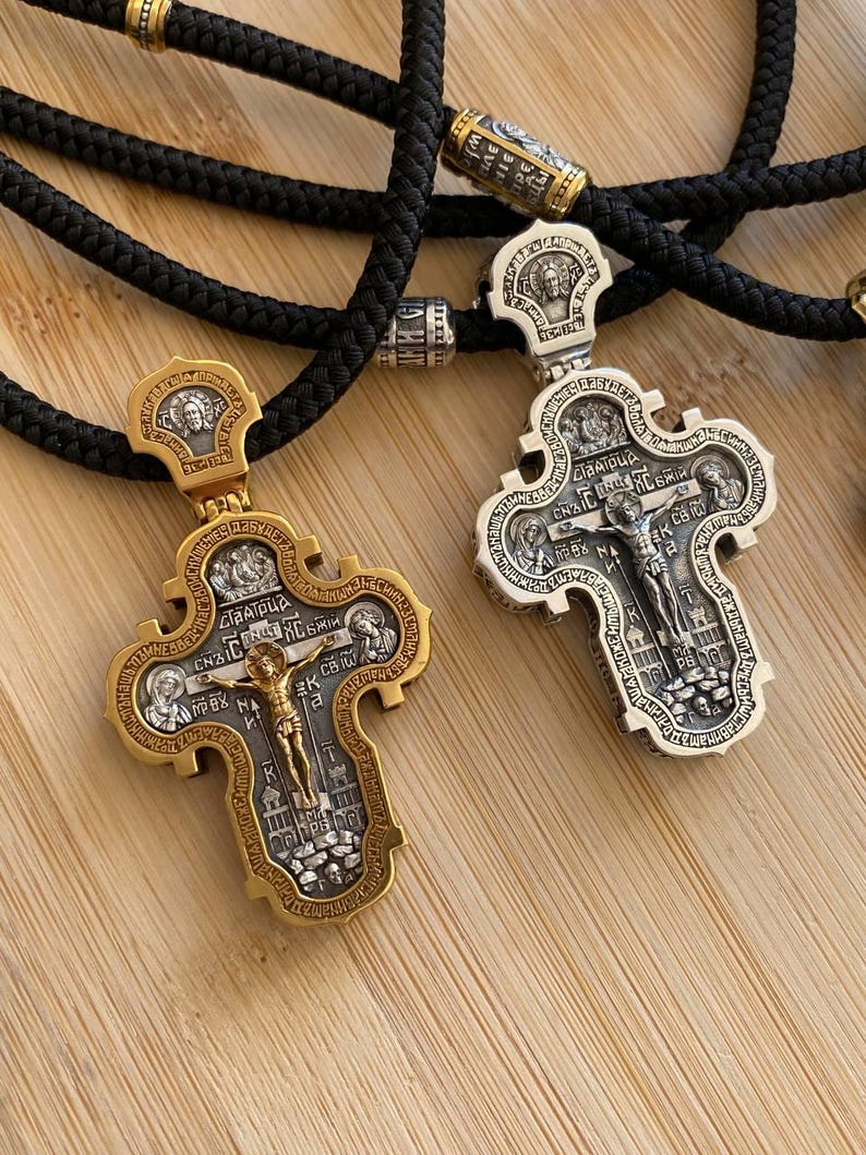 May include: Two ornate religious pendant necklaces. One is gold-toned, the other silver-toned, both with intricate detailing and a central figure. The pendants are suspended from black braided cords. The background is a light wood surface.