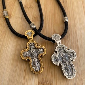 May include: Two ornate religious pendant necklaces. One pendant is gold-toned, the other silver-toned, both with intricate religious figures. The pendants hang from black cords with decorative beads. The background is a light brown wooden surface.