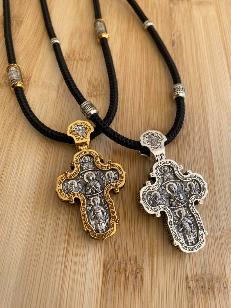 May include: Two ornate religious pendant necklaces. One pendant is gold-toned, the other silver-toned, both with intricate religious figures. The pendants hang from black cords with decorative beads. The background is a light brown wooden surface.