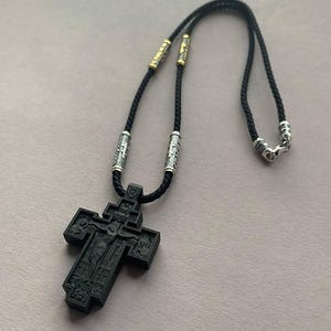May include: A black Orthodox cross pendant necklace. The cross features intricate carvings and is suspended from a black cord with silver and gold-toned accents. The necklace is designed with a clasp for easy wear.