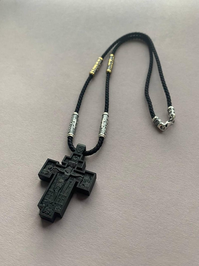 May include: A black Orthodox cross pendant necklace. The cross features intricate carvings and is suspended from a black cord with silver and gold-toned accents. The necklace is designed with a clasp for easy wear.