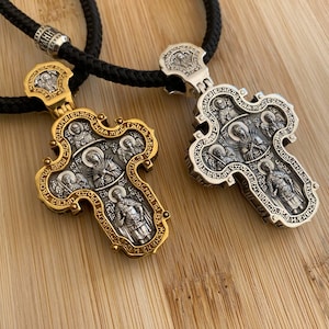 May include: Two ornate religious pendant necklaces. One is gold-toned, the other silver-toned, both with intricate religious figures and text. The pendants are suspended on black braided cords. The background is a light brown wooden surface.