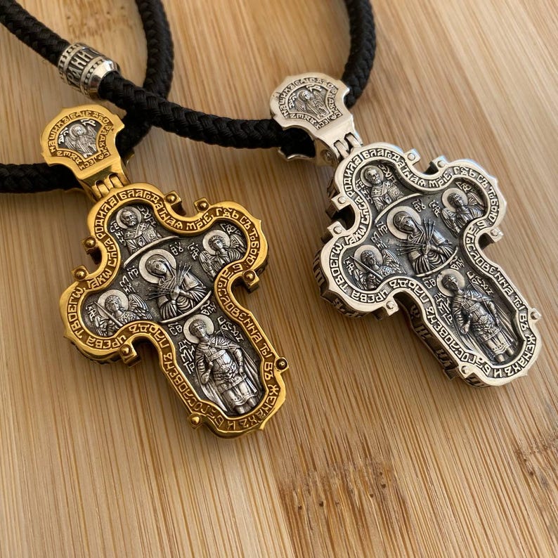 May include: Two ornate religious pendant necklaces. One is gold-toned, the other silver-toned, both with intricate religious figures and text. The pendants are suspended on black braided cords. The background is a light brown wooden surface.