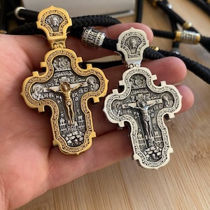 May include: Two ornate, gold and silver religious crosses with intricate detailing, including figures and text. The crosses are suspended on black cords, showcasing their detailed craftsmanship and religious significance.