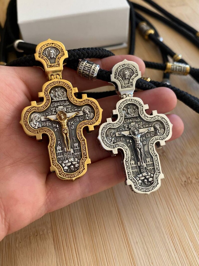 May include: Two ornate, gold and silver religious crosses with intricate detailing, including figures and text. The crosses are suspended on black cords, showcasing their detailed craftsmanship and religious significance.
