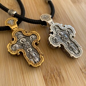 May include: Two ornate religious pendant necklaces. One is gold-toned, the other silver-toned. Both feature detailed religious figures and text, suspended from black cords. The pendants are on a wooden surface.