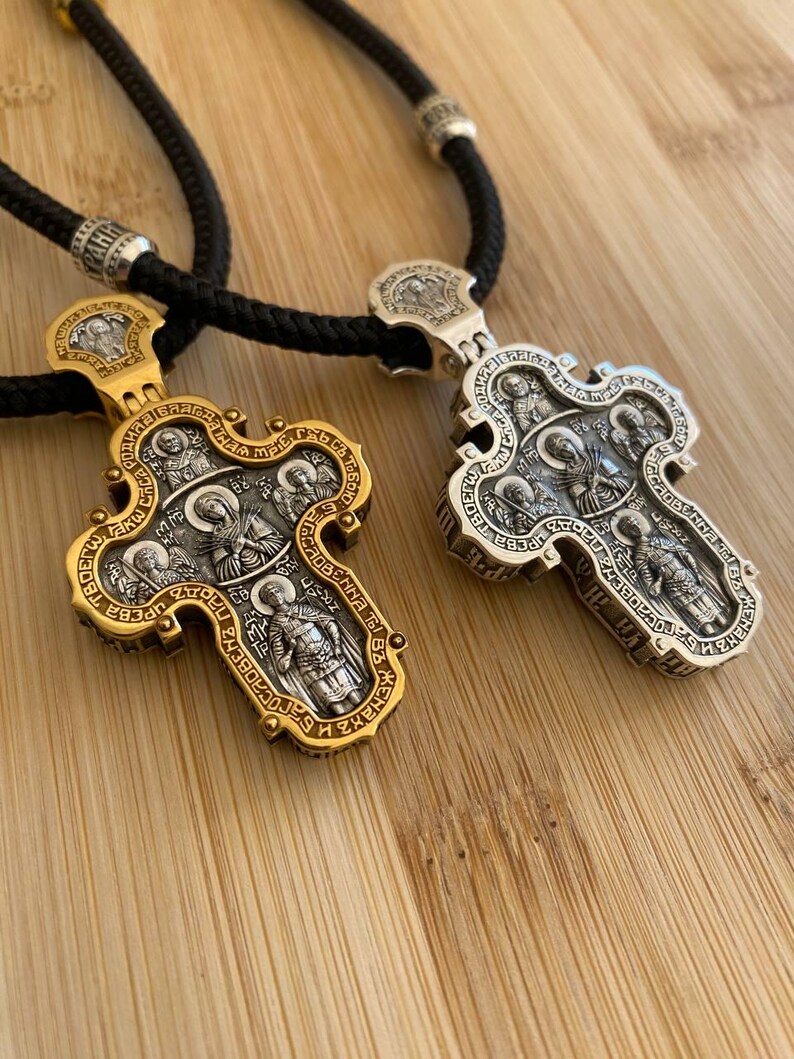 May include: Two ornate religious pendant necklaces. One is gold-toned, the other silver-toned. Both feature detailed religious figures and text, suspended from black cords. The pendants are on a wooden surface.