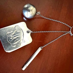 May include: A silver-toned chatelaine accessory featuring a rectangular engraved tag with ornate script initials, a spherical container, and a cylindrical tube, all connected by delicate metal chains to a central circular ring.
