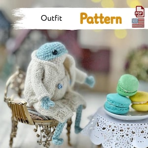 May include: A crochet frog figure with a light blue head and limbs, wearing a beige knitted coat with small blue buttons. The frog sits on a gold-toned ornate chair next to a small table with a white lace cover and three colorful macarons.