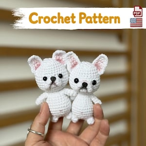 May include: A digital PDF crochet pattern featuring two small, white, handmade amigurumi French bulldog figures with black plastic eyes, black noses, and pink-tinted ears. The text Crochet Pattern is displayed at the top of the image.