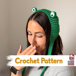 May include: A bright green crochet hat featuring two protruding frog eyes with white and black pupils on top. The hat has long side flaps and is labeled with the text Crochet Pattern and a small United States flag icon in the corner.