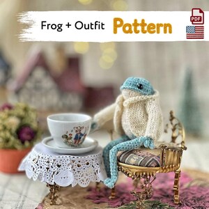 Könnte beinhalten: Ein gehäkelter Frosch in einer cremefarbenen Strickjacke, sitzend auf einem kleinen goldfarbenen Stuhl neben einer weißen Teetasse mit Blumenmuster auf einem mit weißer Spitze bedeckten Tisch. Das Bild enthält den Text Frog + Outfit Pattern und ein PDF-Symbol.