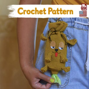 Peut inclure: Une pochette en crochet jaune moutarde en forme de chien avec un cordon de serrage, des yeux en feutrine, un nez noir et une langue rouge. La pochette possède une ouverture en bas pour distribuer des sacs. Le texte Crochet Pattern est visible.