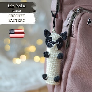 Puede incluir: Patrón de crochet para un estuche de bálsamo labial con forma de perro blanco y negro. El estuche está unido a una bolsa rosa con cremallera.