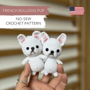Peut inclure: Deux chiots bouledogues français au crochet blancs, avec des yeux et des nez noirs, et l'intérieur des oreilles rose. Le texte "FRENCH BULLDOG PUP" et "NO-SEW CROCHET PATTERN" est inclus. Un drapeau américain est dans le coin supérieur droit.