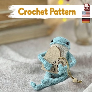 May include: A light blue crocheted frog figurine with black bead eyes and a white belly, holding a wooden heart-shaped cutout with intricate laser-cut details. The text Crochet Pattern is displayed at the top with US and German flag icons.