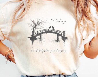 Può includere: Una maglietta bianca con una grafica in bianco e nero di un ponte con due uccelli sopra. Il testo "Love is the bridge between you and everything" è stampato sotto la grafica.