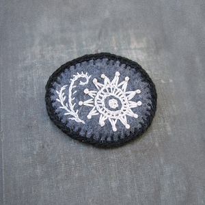 May include: A black felt brooch with a white embroidered sun and a small white embroidered stem.