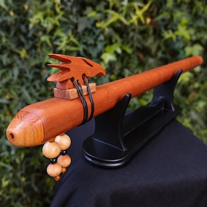 May include: A long, cylindrical wooden flute with a reddish-brown finish. It features a carved bird-shaped block secured by black cords on top. A cluster of light-colored wooden beads hangs from the side. The flute rests on a black stand.