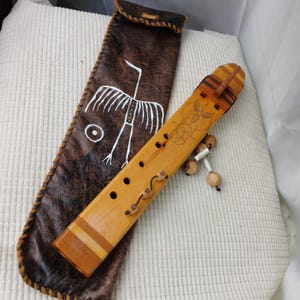 May include: A wooden flute with a decorative design and a leather case. The flute is light brown with dark brown accents and several finger holes. The case is dark brown with a white bird design and a braided border.