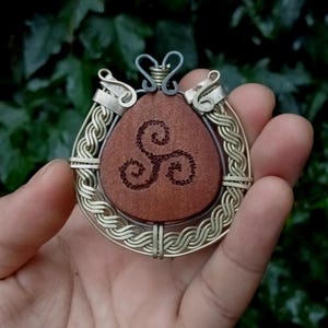 Puede incluir: Un colgante con un centro de madera marrón triangular y un símbolo de trisquel marrón oscuro grabado en el medio. La madera está encerrada en un marco de alambre plateado con intrincados patrones trenzados y un lazo decorativo superior.