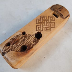 Puede incluir: Una flauta de madera con un diseño geométrico tallado en la superficie. La flauta tiene seis agujeros y una boquilla en un extremo.
